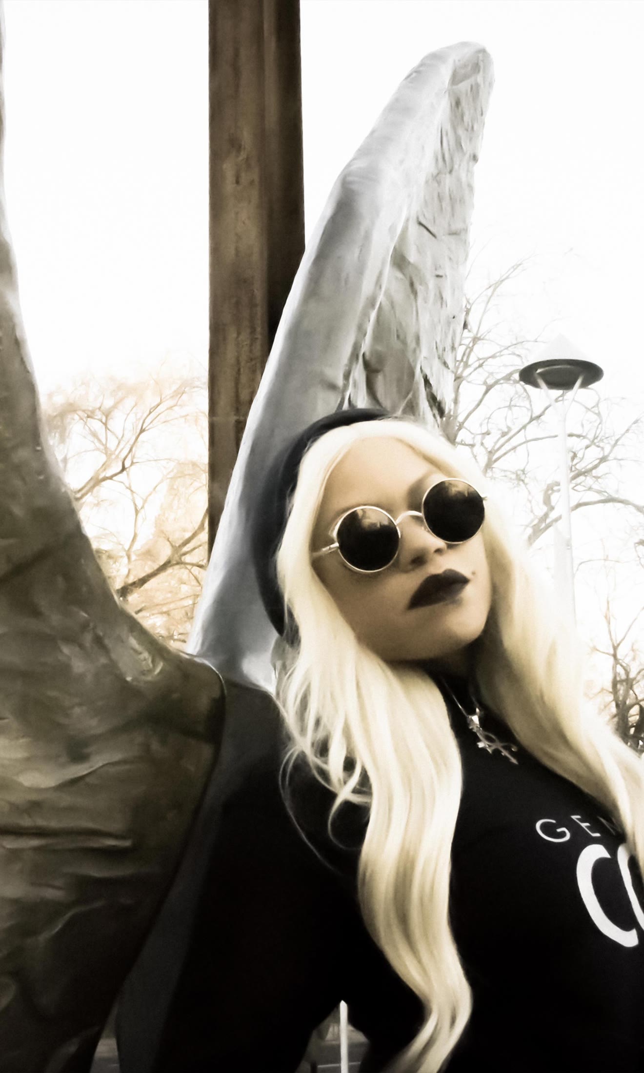Pandora Gunn in round sunglasses and a black beanie, platinum hair, beside a sculpted angel wing